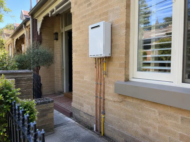 Wall-mounted hot water unit with copper pipework on a brick house exterior beside a window in Paddington